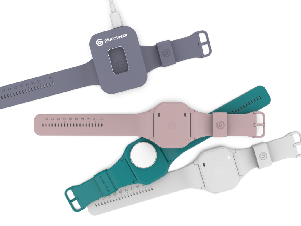 Four Glucowear™ devices shown, watch-like devices with no watch face. The colours of the devices are white, teal, baby pink and mauve. The mauve device is sat in a mauve coloured charger. The teal device is upside down, showing the side that sits on your wrist. It has a round white circle which is the antenna that monitors your blood glucose levels.