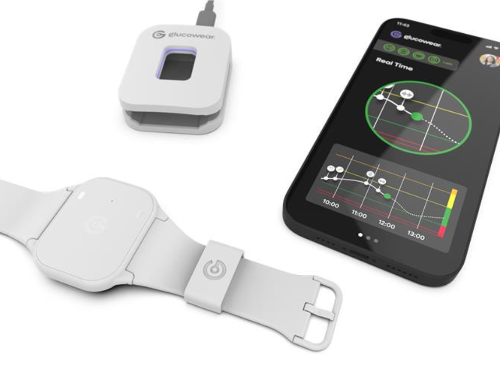 “Glucowear™ device shown a white coloured watch-like device with no watch face. It has a smartphone next to showing an example of the real time data from the Glucowear™ app. There is also a white coloured charger which the device can slot into to be charged.