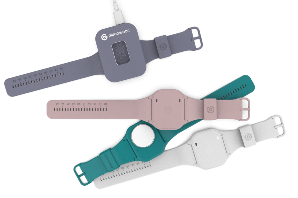Four Glucowear™ devices shown, watch-like devices with no watch face. The colours of the devices are white, teal, baby pink and mauve. The mauve device is sat in a mauve coloured charger. The teal device is upside down, showing the side that sits on your wrist. It has a round white circle which is the antenna that monitors your blood glucose levels.