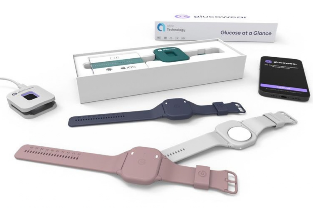 Assorted wearable Glucowear™ devices laid out on a white background, with different colour bands, a small black monitor, and a charging base in a product box.